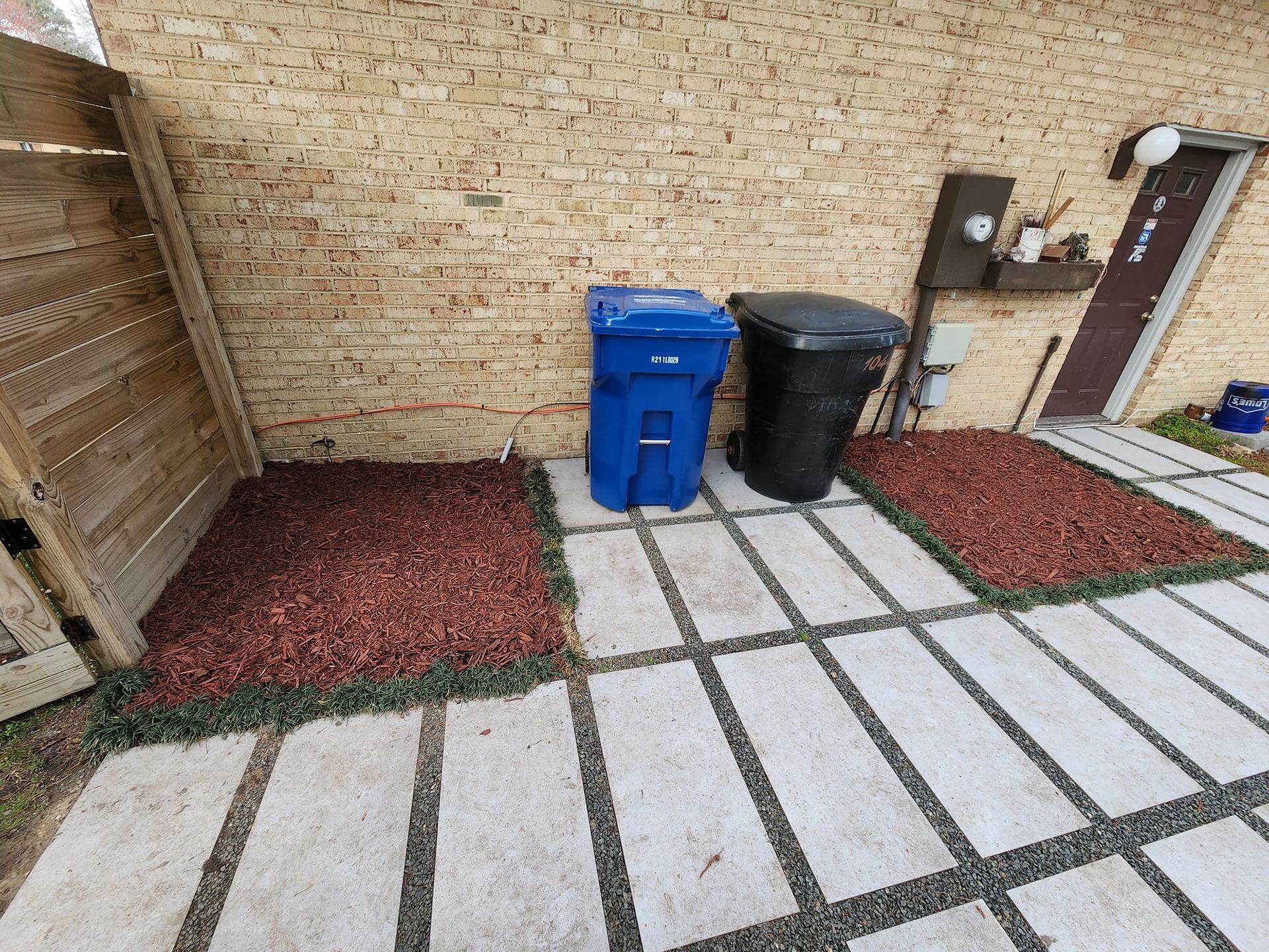 A backyard with concrete pavers, red gravel, two trash bins, and a wooden fence.