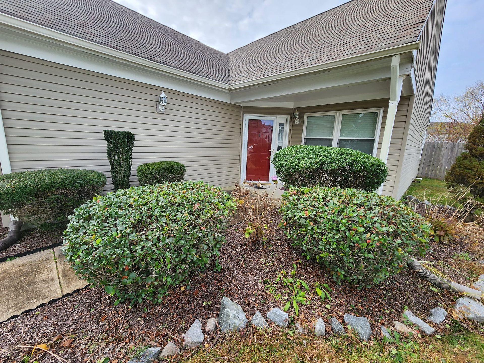 A house exterior with brown siding, a red door, and green bushes in the front yard.