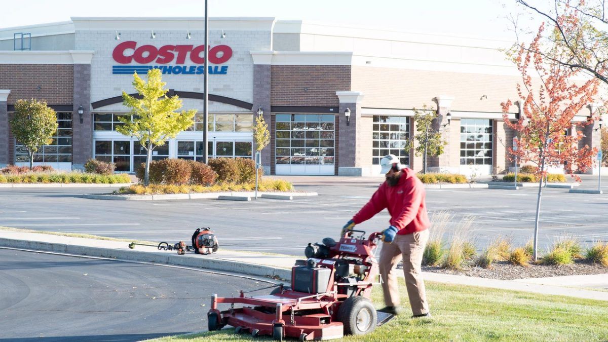 Man mowing the lawn in front of a Costco store. Red lawnmower, person in red shirt. Outdoors.