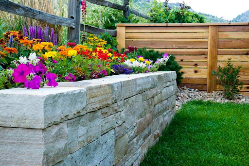 Stone planter box overflowing with colorful flowers next to wooden fence and green grass.