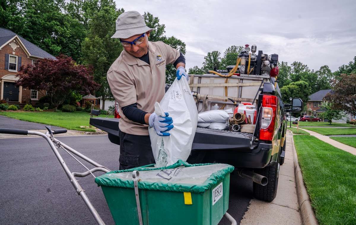 Man in hat and gloves pouring from a bag into a spreader, near a truck.