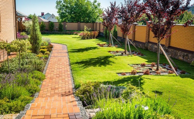 Brick pathway through a lush green lawn, lined with plants and trees, next to a brick fence.