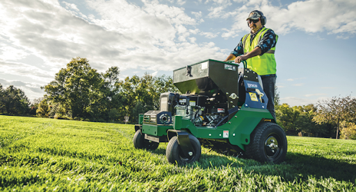 Man operating a green aerator on a grassy field, wearing safety gear under a bright sky.