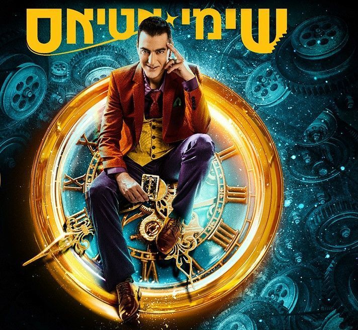 גבר מצולם בין ידיים מורמות, מביט למצלמה על רקע כהה.