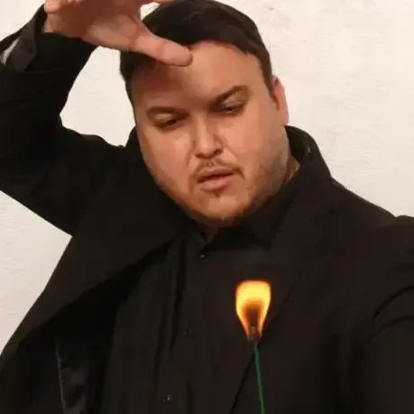 אדם בחליפה שחורה בתנוחה דרמטית, ידו על מצחו; אפקט להבות באזור החזה.