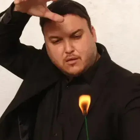 גבר בחליפה שחורה, שמחזיק יד מעל ראשו, ולהבה קטנה סמוך לחזהו.