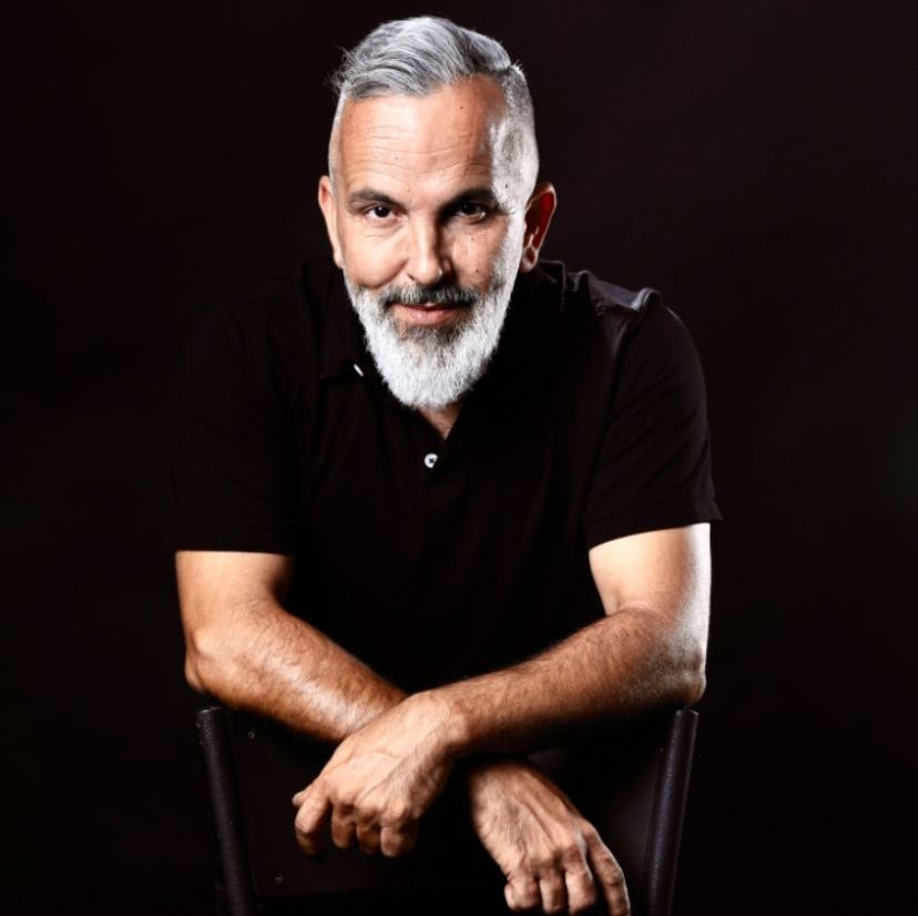 דיוקן סטודיו של גבר עם שיער אפור וזקן, לבוש בחולצה שחורה על רקע כהה