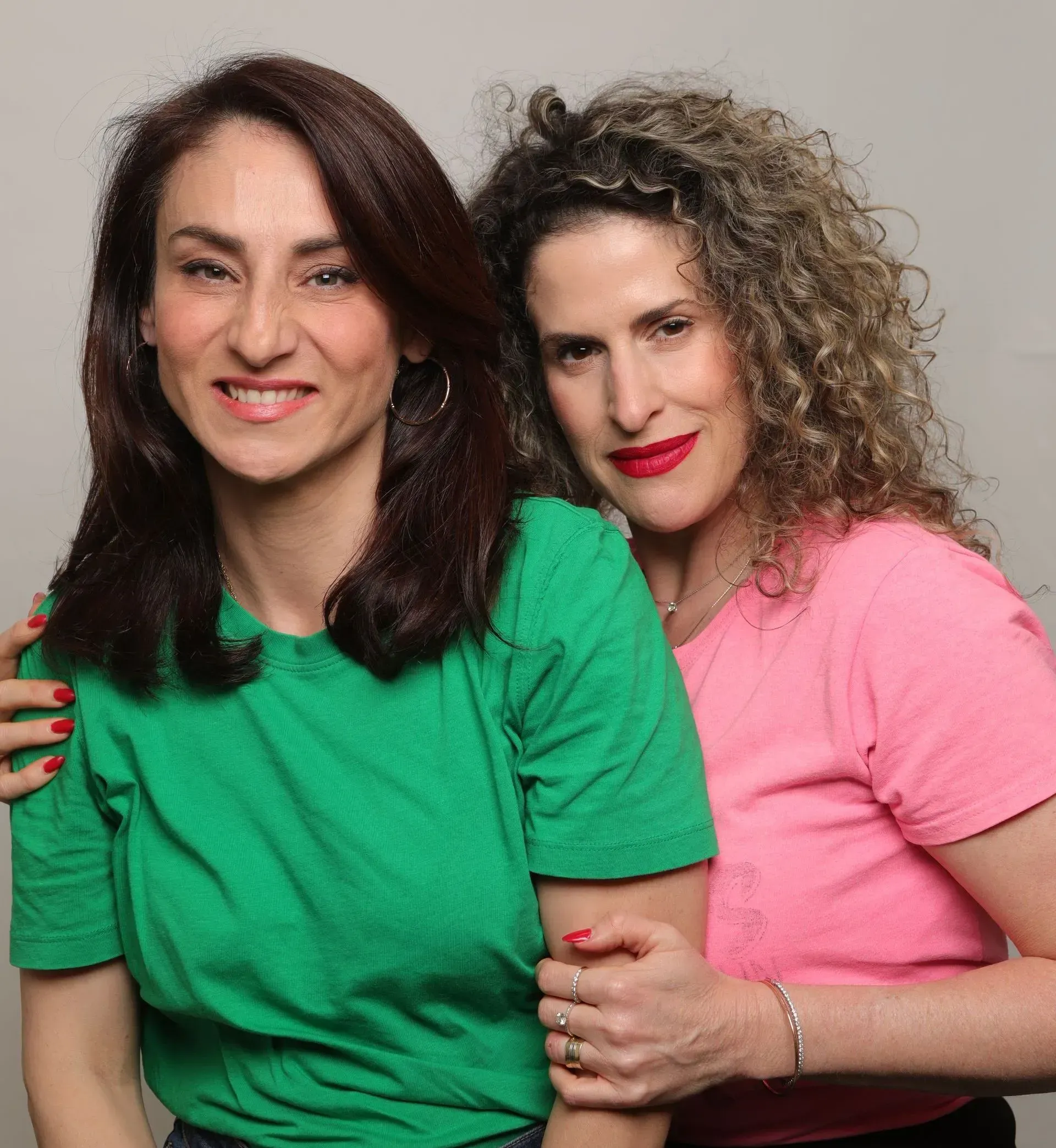 שתי נשים מצטלמות יחד, אחת בחולצה ירוקה והשנייה בחולצה ורודה, ומחייכות על רקע חלק