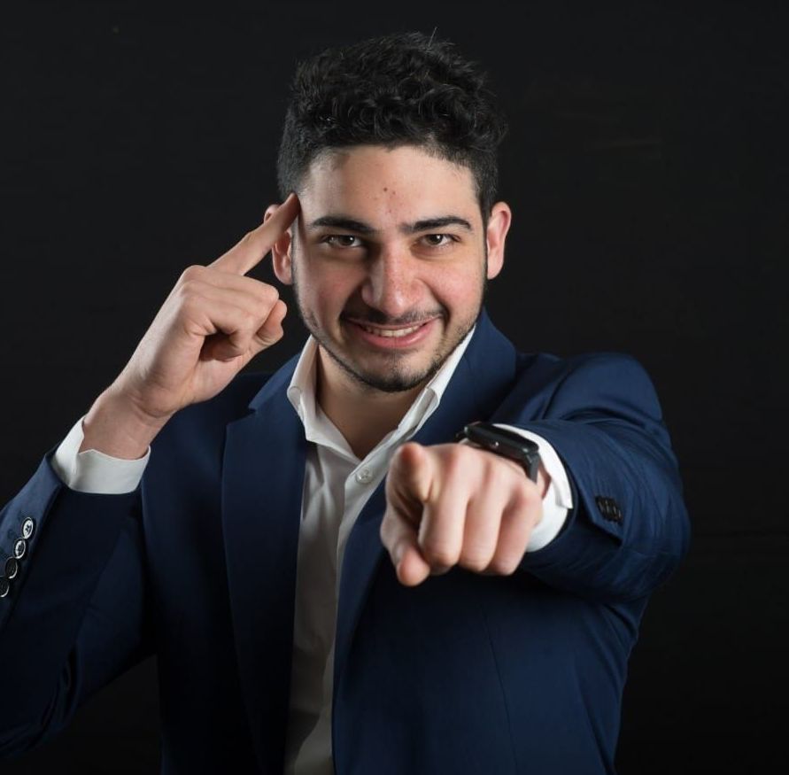 גבר בחליפה כחולה כהה מצביע על הצופה ומקיש על רקתו על רקע כהה.