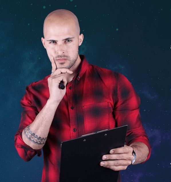 גבר קירח בחולצה משובצת אדומה, מחזיק לוח כתיבה ומניח את ידו על סנטרו על רקע כהה