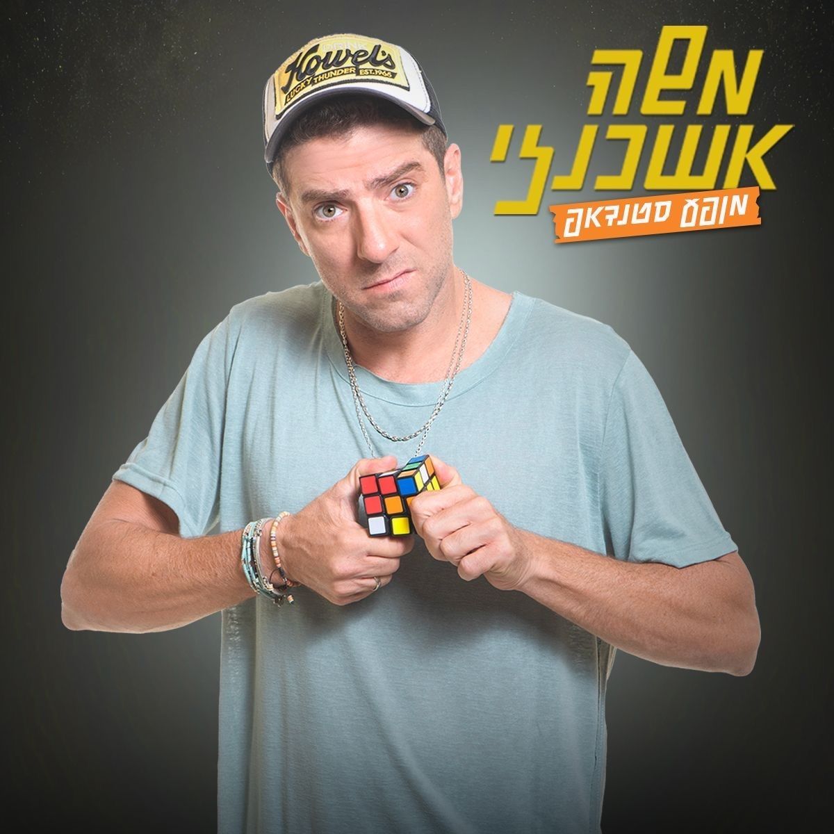 גבר מחזיק בקוביית רוביק, לובש חולצה וכובע בצבע תכלת, לצד טקסט בעברית בצהוב על רקע אפור