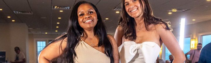Two smiling women in light-colored dresses at an indoor event with warm lighting.
