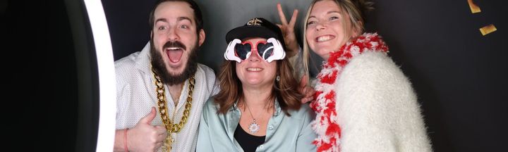 Three smiling people posing in colorful feather boas and party accessories in a photo booth