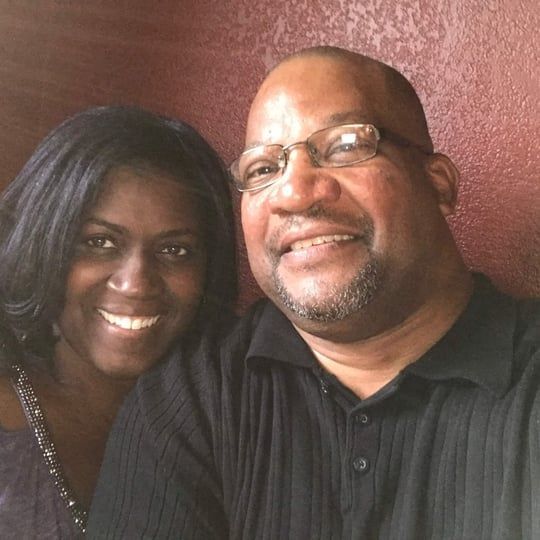 Two smiling people posing closely together indoors against a reddish wall.