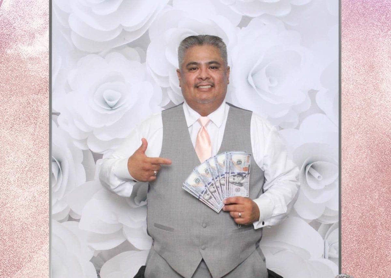 Smiling man in a gray vest holding cash fan in front of white floral backdrop
