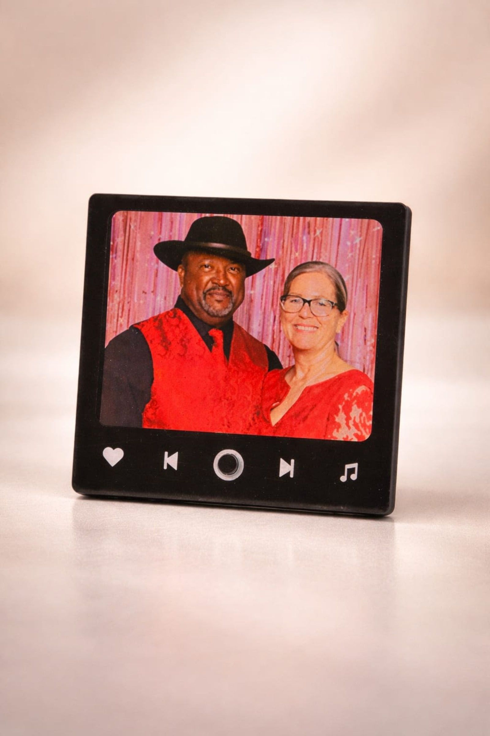 Small digital photo frame showing two people posing against a pink backdrop, with heart and music icons below.