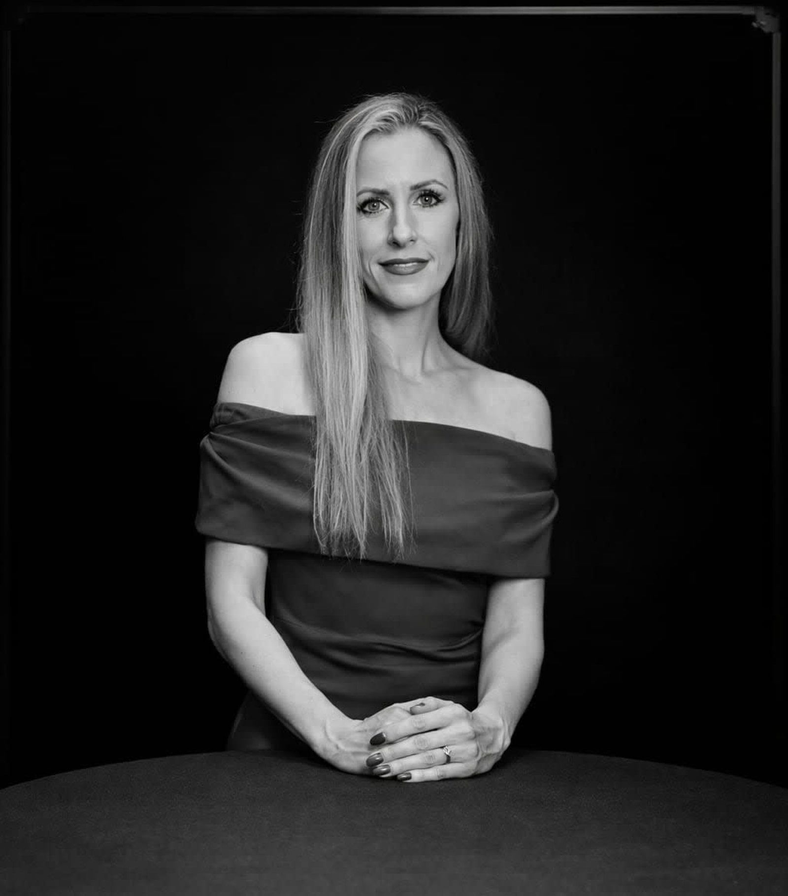 Black-and-white portrait of a woman seated at a table against a dark background, wearing an off-the-shoulder dress.