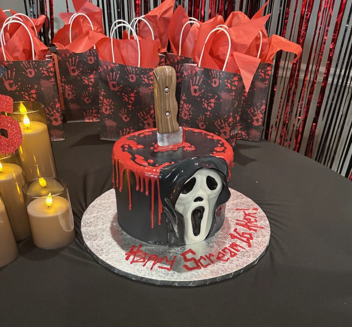 Halloween cake with red dripping icing and Ghostface face on a black table, with red gift bags behind