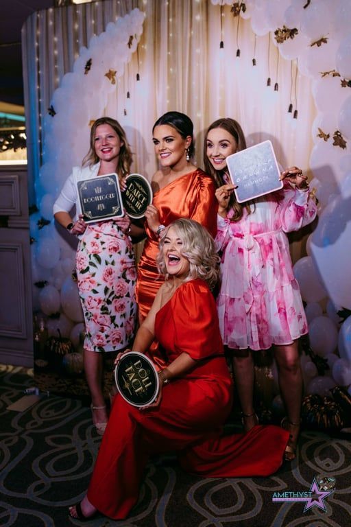 Four women in colorful dresses pose with awards at a formal event backdrop.