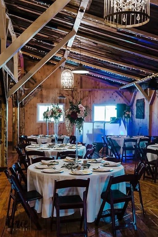 Rustic banquet hall with round tables, black chairs, white tablecloths, and string lights under wooden beams