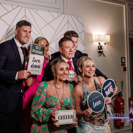 Group of people posing at a party, holding novelty signs and smiling in front of a decorated wall.