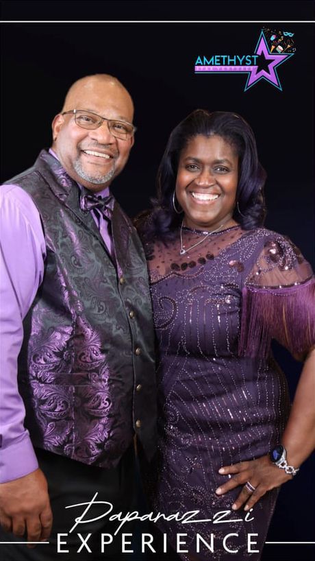 Smiling couple in purple outfits posing against a black backdrop at an experience event