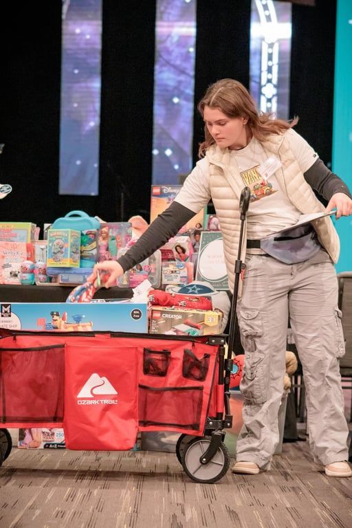 Woman placing items into a red cart beside a blue table at an indoor event