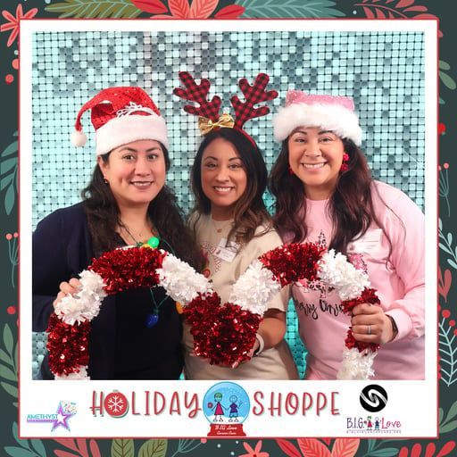 Three women in festive hats pose behind red-and-white holiday decorations at a Holiday Shoppe photo backdrop.