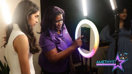 Three people at a makeup station, one using a ring light to adjust a phone or mirror.