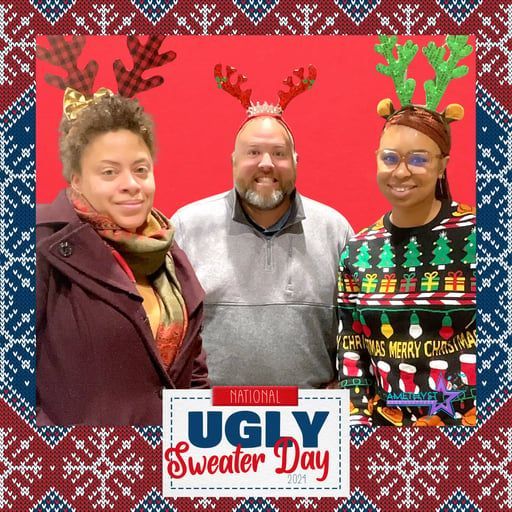 Three people posing in festive holiday sweaters and antler headbands before a red “Ugly Sweater Day” backdrop.