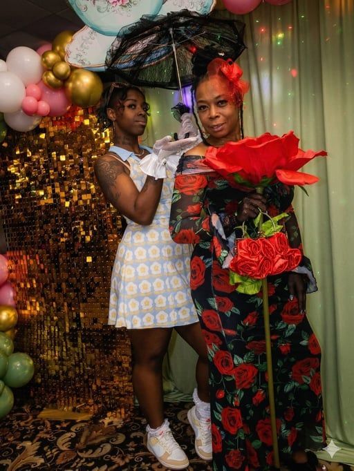 Two people posing at a festive balloon backdrop, holding large red flowers and wearing patterned outfits