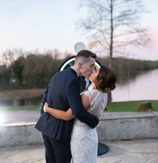 Couple kissing by a waterfront at sunset, the woman in a white lace dress and the man in a dark suit