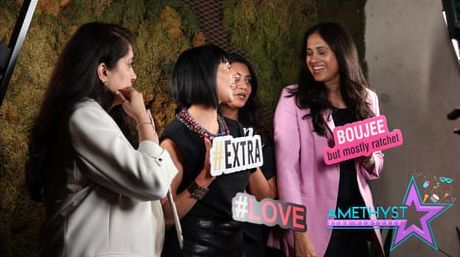 Four women chatting indoors with colorful labels like “EXTRA,” “LOVE,” and “BOUJEE” around them.