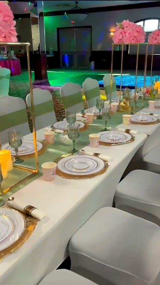 Decorated banquet table with white chairs, gold accents, and pink floral centerpieces in a colorful event hall