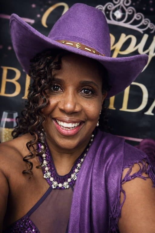 Smiling person in a purple cowboy hat and dress at a celebratory backdrop