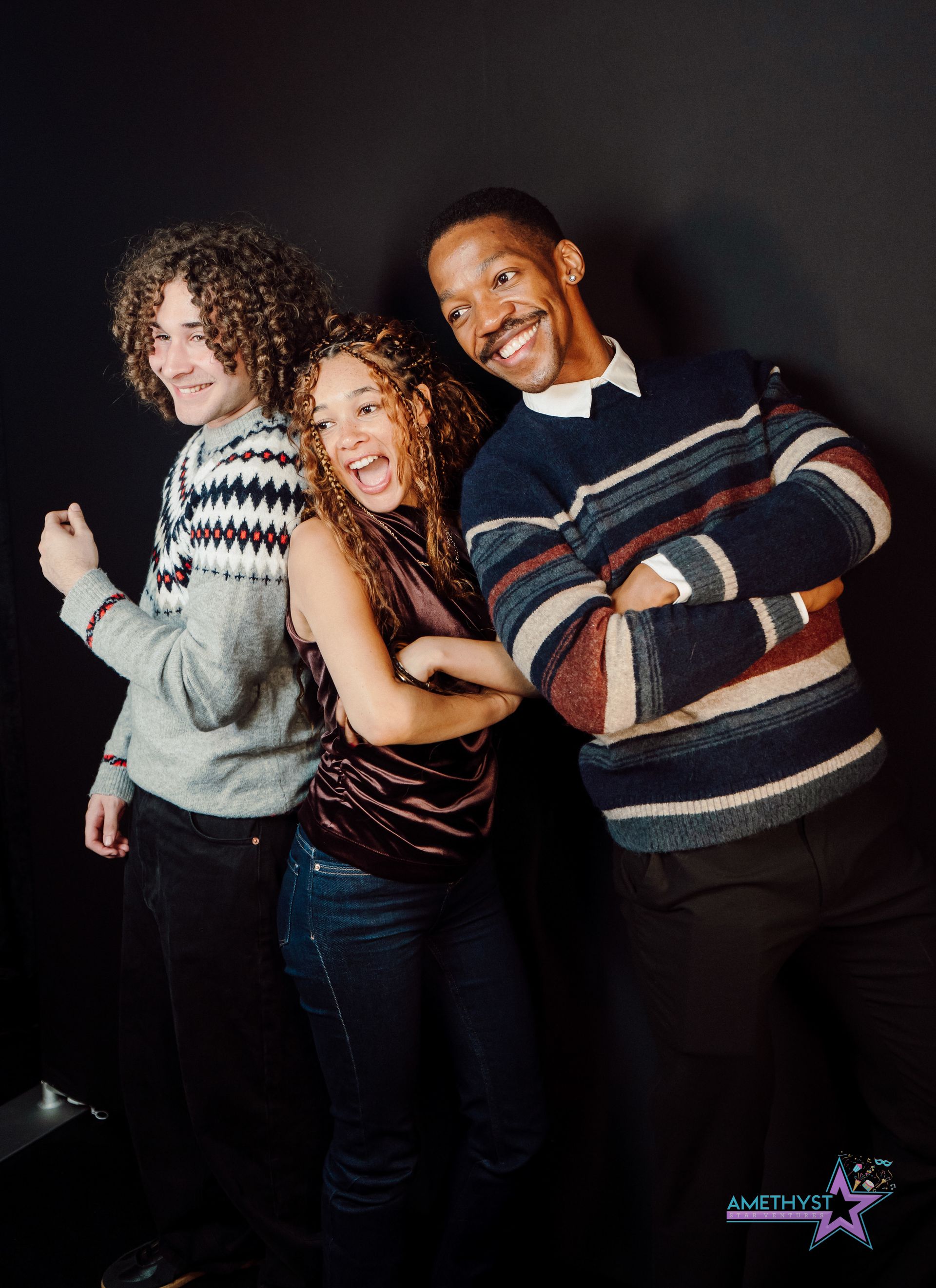 Three people smiling and posing together against a dark studio backdrop, wearing cozy sweaters.