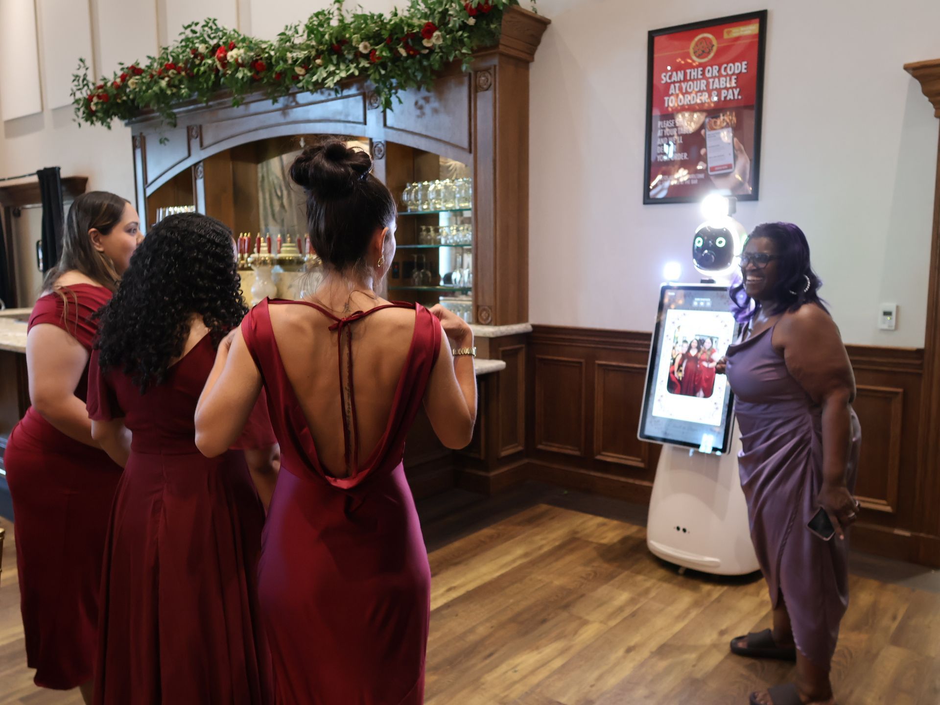 Four wedding guests in burgundy dresses reviewing a photo on Starla's touchscreen with an Event Ambassador
