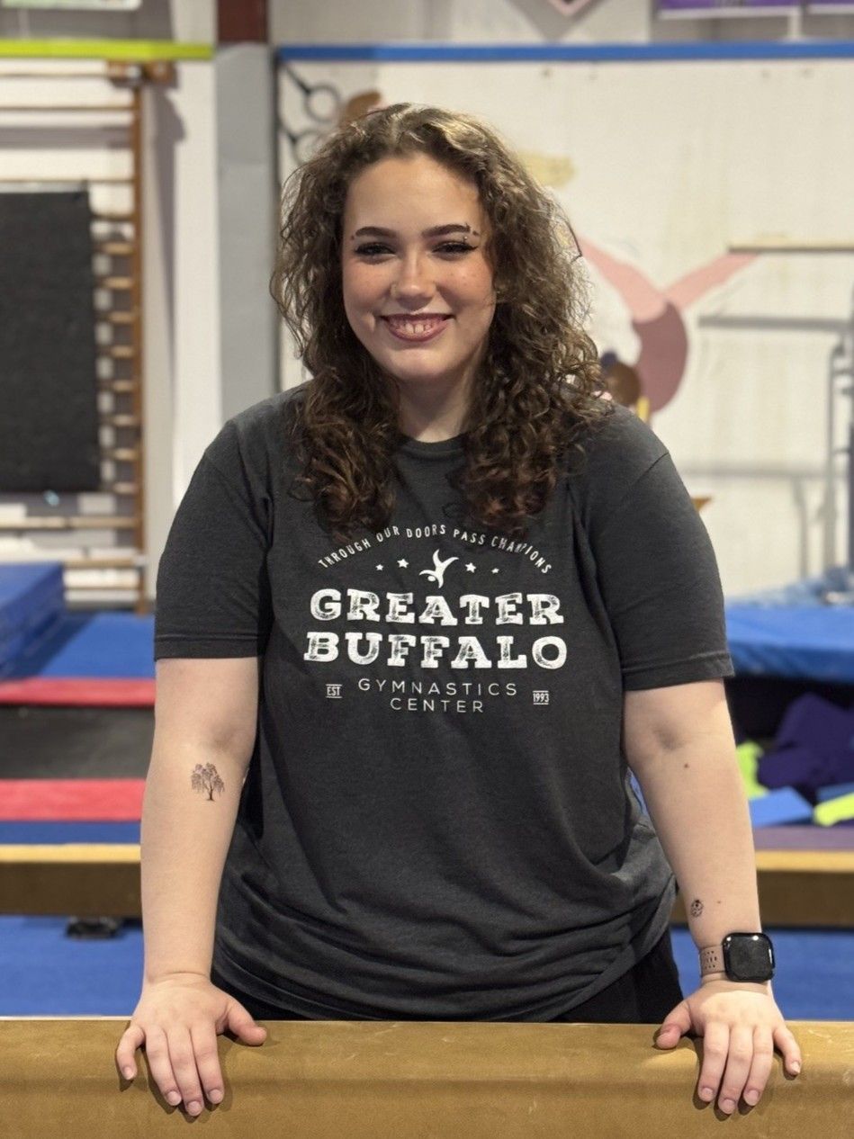 A woman wearing glasses and a black polo shirt is standing in a gym with her hands on her hips.