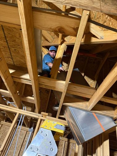 Worker in blue hard hat standing in a wooden attic framing area, viewed from below.