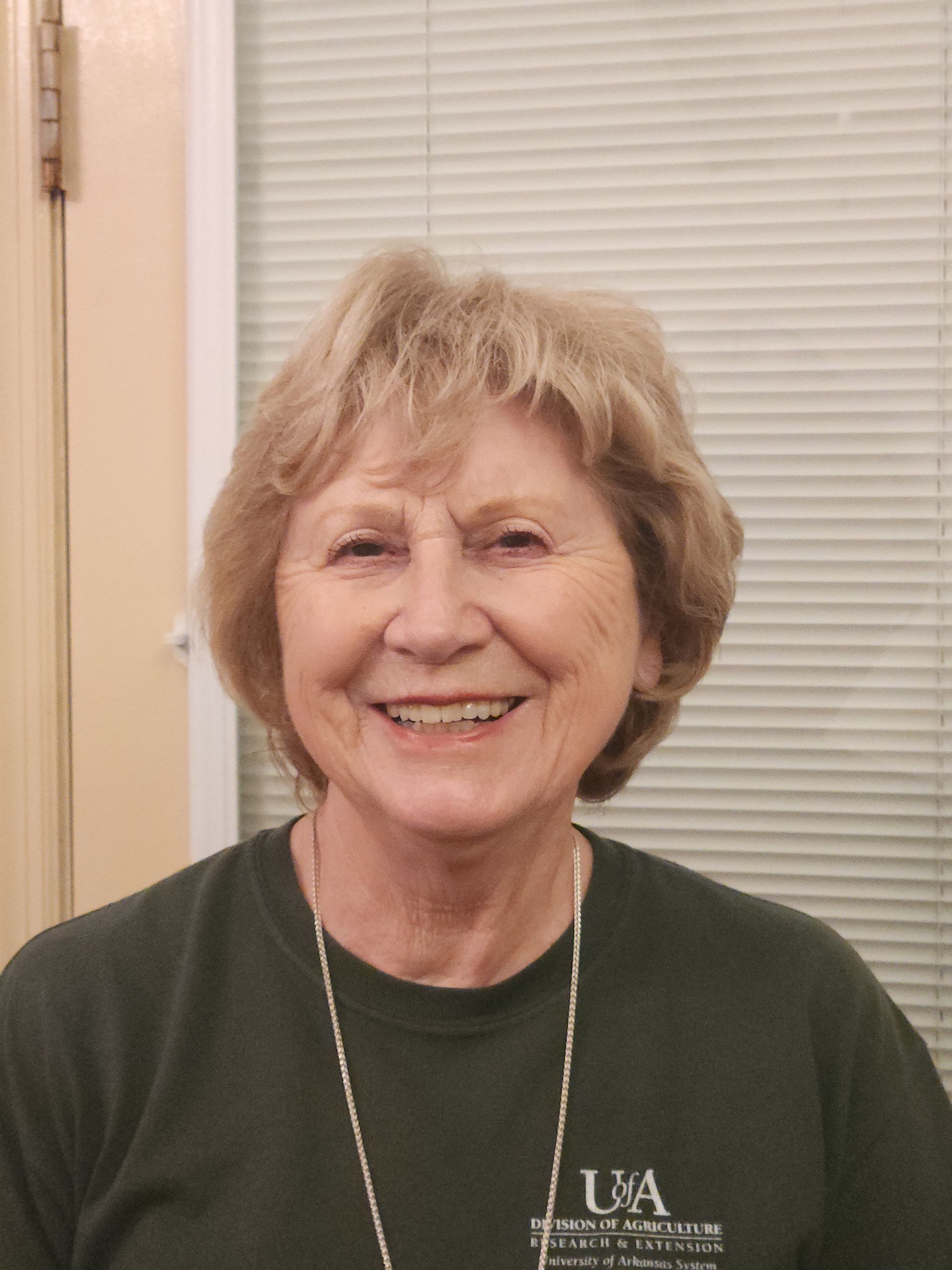 A smiling person with shoulder-length blonde hair, wearing a green t-shirt with a UVA logo, stands before window blinds.