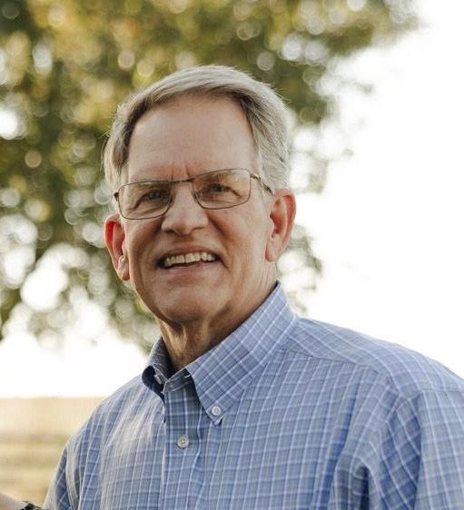 A man with glasses and grey hair wears a blue plaid button-down shirt, smiling outdoors with a blurred tree in the back.