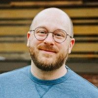 A person with a shaved head and a beard wearing round-framed glasses, smiling against a wood-paneled background.