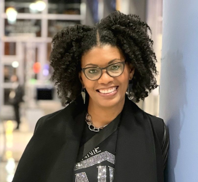 Smiling woman with curly hair and glasses, wearing a dark blazer and necklace, leaning against a blue wall.