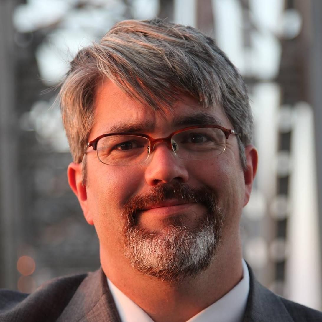 A person with grey-streaked hair and glasses, wearing a suit and collared shirt, looks toward the camera.