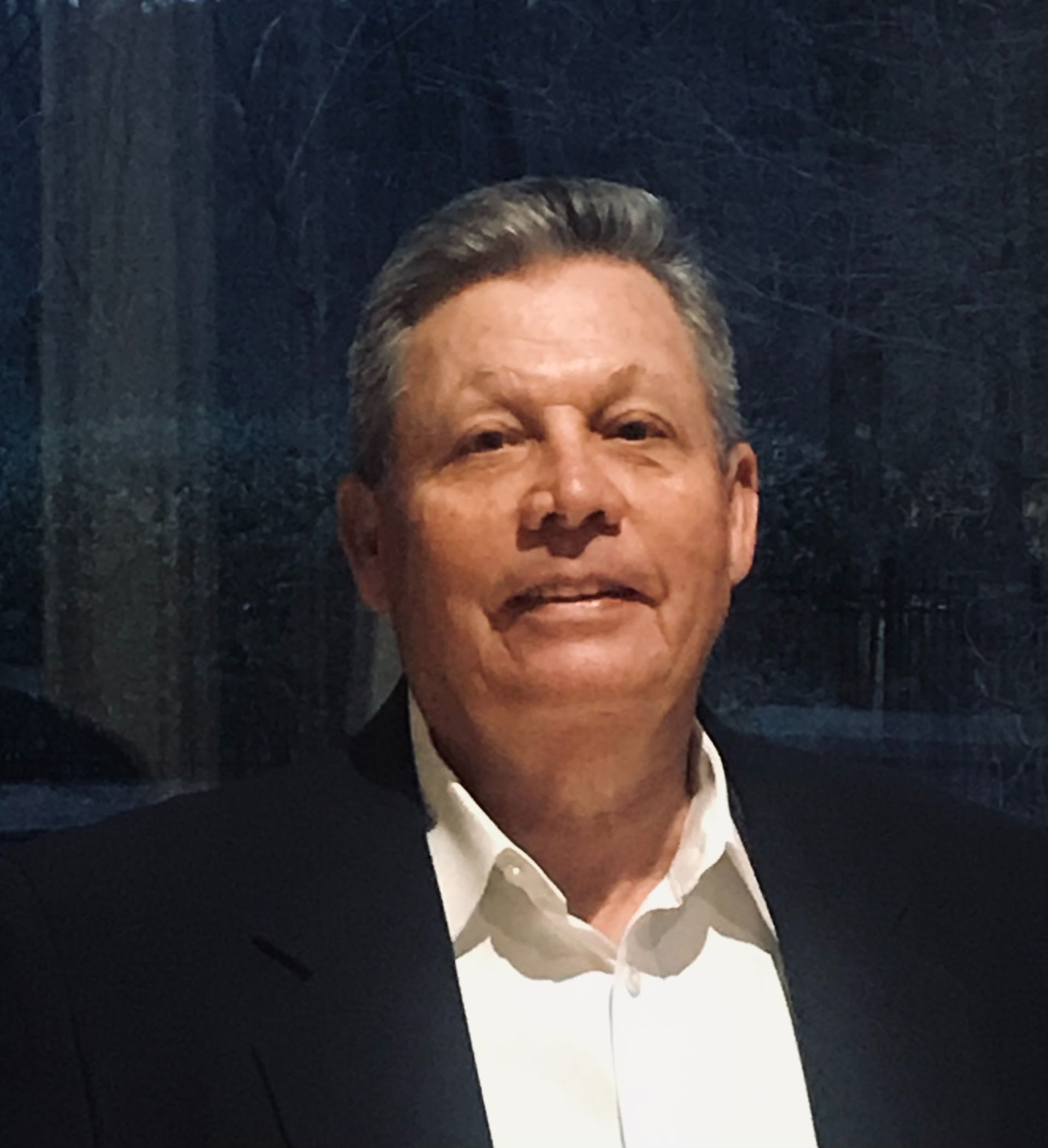 A person with gray hair wearing a white dress shirt and a black blazer, posed in front of a window.
