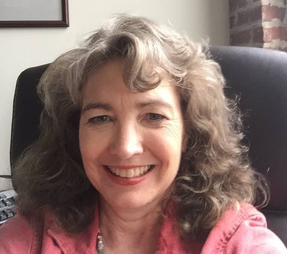 A person with shoulder-length, wavy hair smiles at the camera, wearing a pink shirt, seated in an office chair.