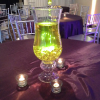 Glowing green drink in a glass centerpiece on a purple-lit table with small candles at a formal event