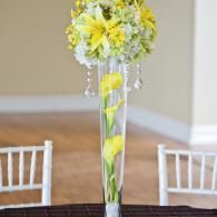 Tall clear vase with yellow and white flowers and hanging crystals on a table at an indoor event