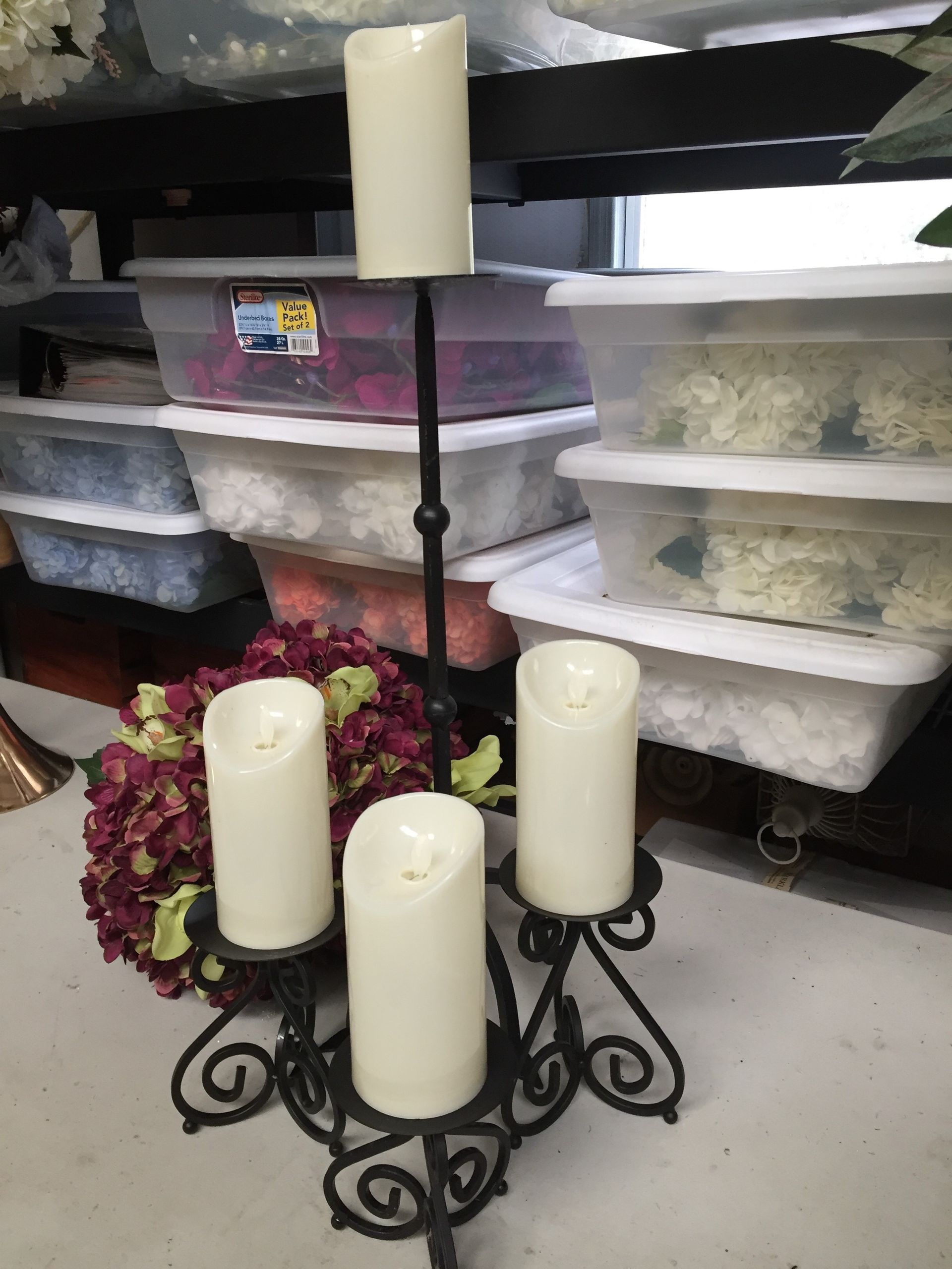 Black wrought-iron candle holder with four white candles, in front of stacked storage bins with flowers