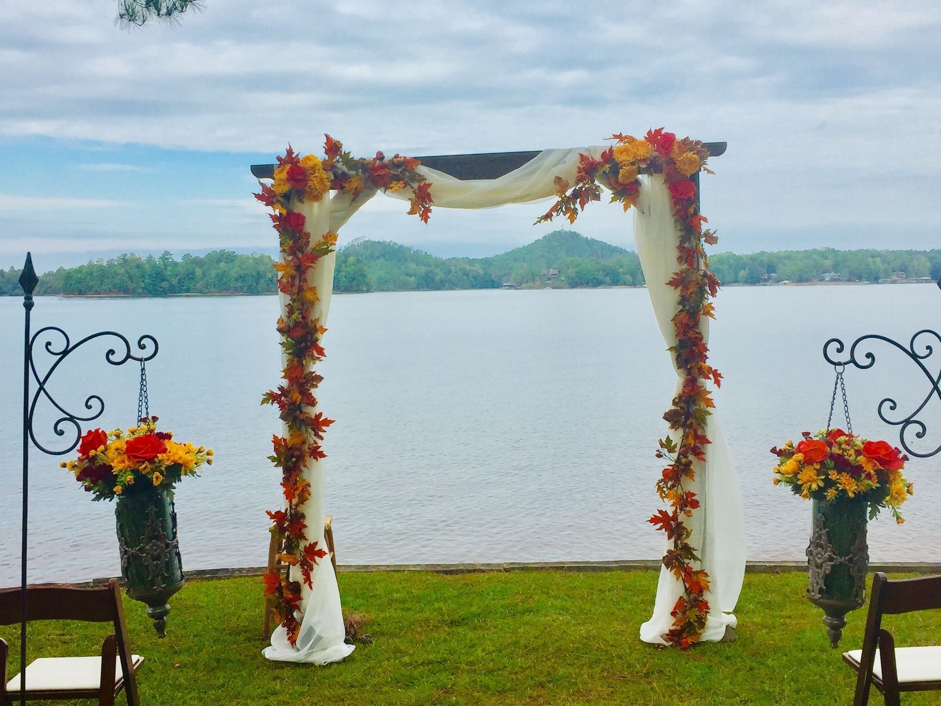 Autumn floral wedding arch on a lakeside lawn with decorative lantern stands and mountains beyond